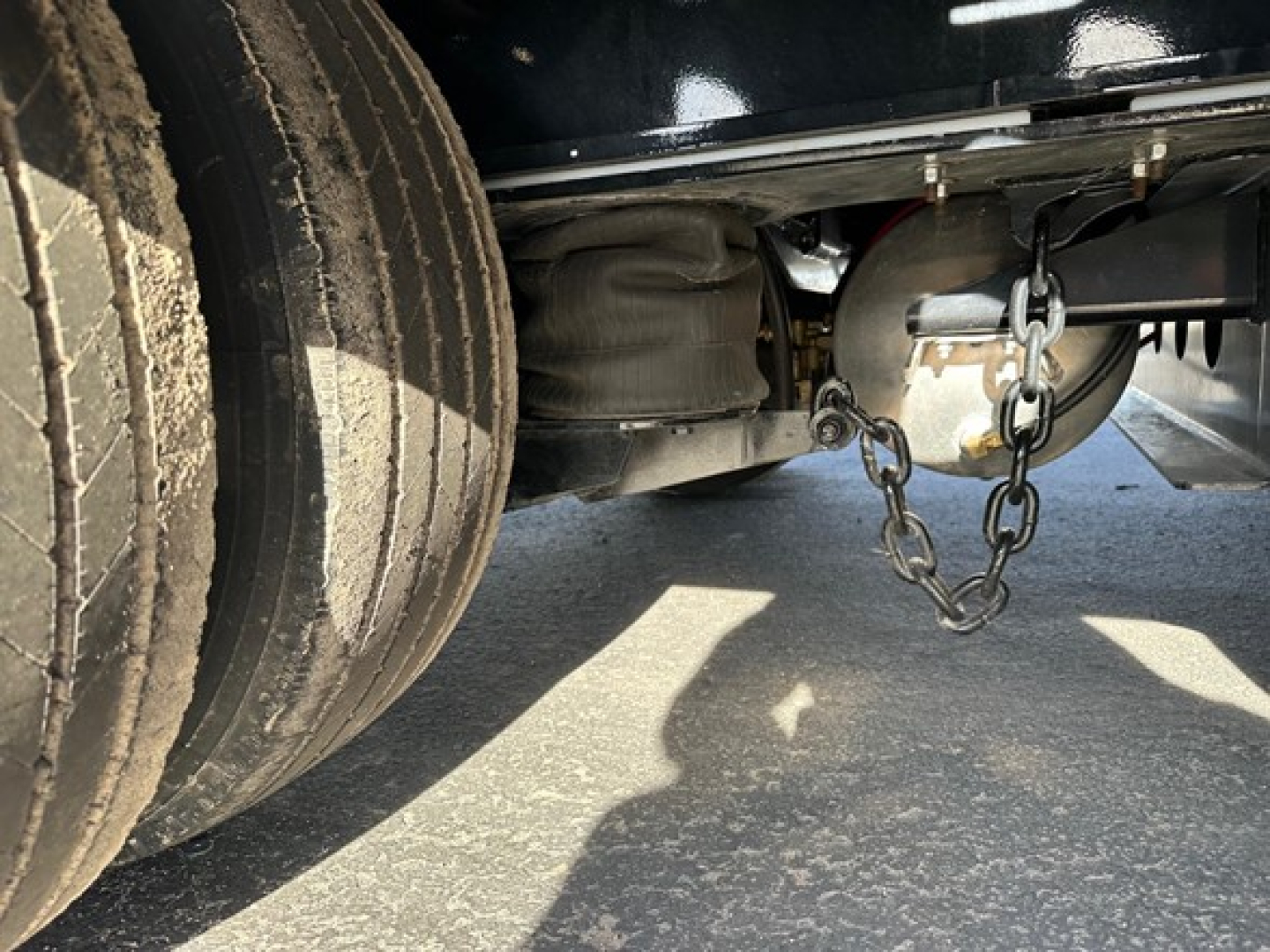 Trailer undercarriage showing axles, dual tires, and a steel chain hitch with a wheel hub and suspension components.