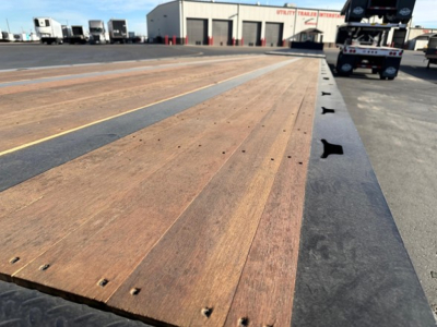 Flatbed trailer deck with wooden planks and black steel edge, viewed from low angle; Doepker tridem RT trailer yard in background.