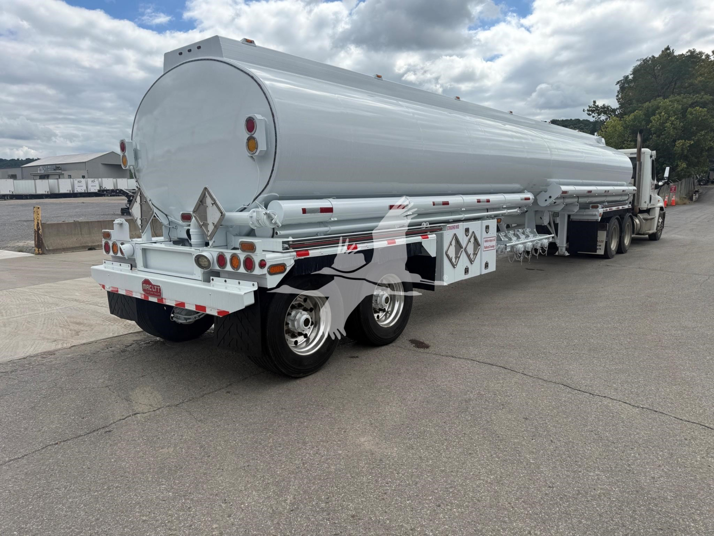 Gasoline tanker trailer with cylindrical tank and rear dual axle, viewed from the left-rear; light gray paint, safety reflectors, rear lights, and warning placards visible.