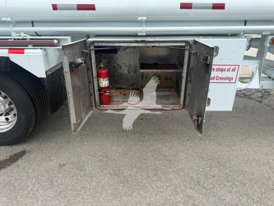 Fuel trailer undercarriage hatch open showing fire extinguisher mounted inside front of storage compartment, with metal interior and warning label on the right side.