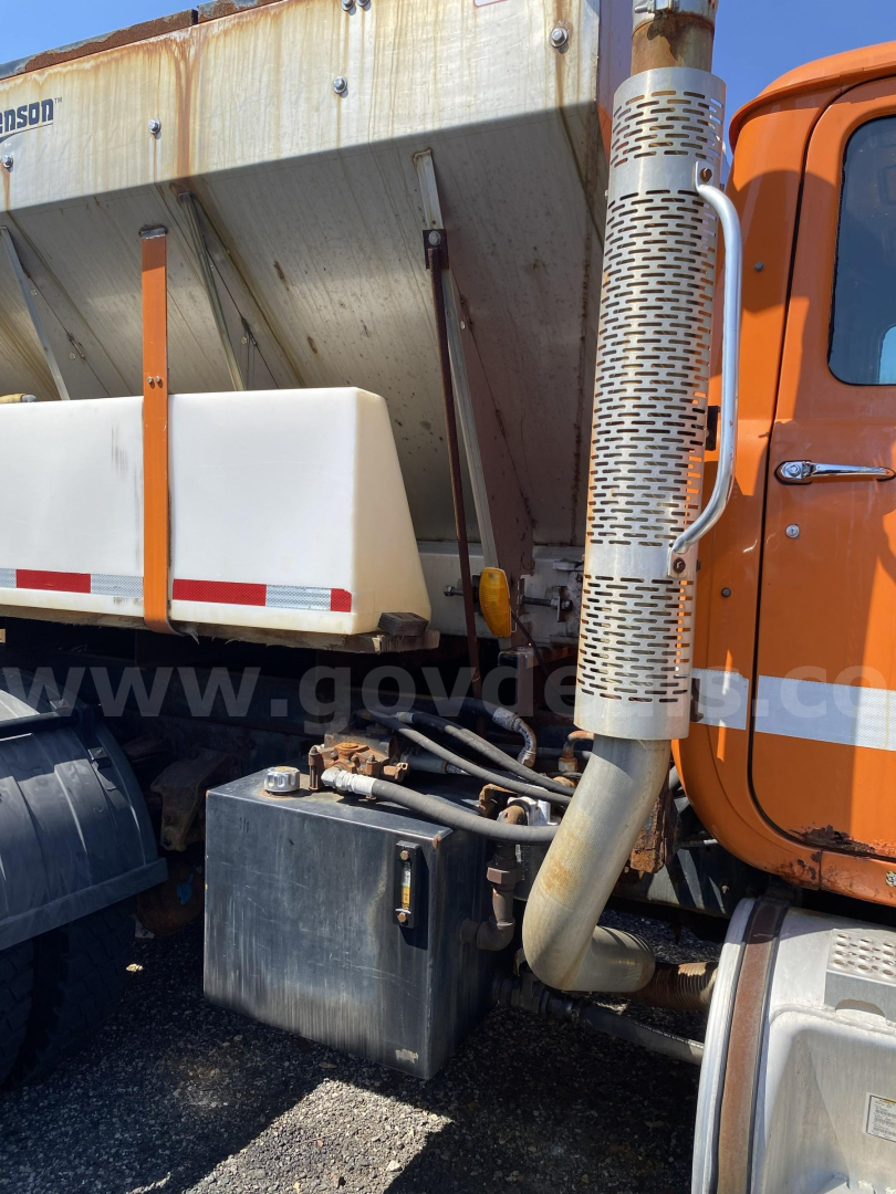 Truck side view showing fuel tank and exhaust stack near cab of orange Mack RD688P plow truck.