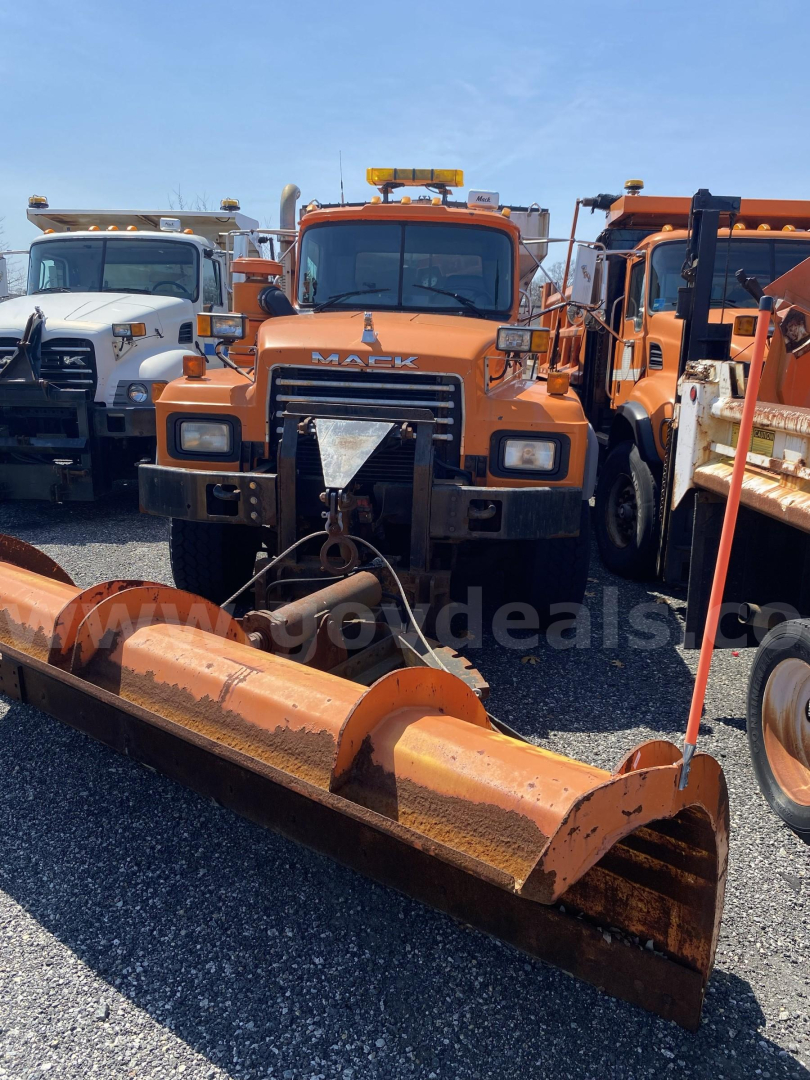 Orange 2004 Mack RD688P plow truck in a row of equipment, with a large metal snow plow blade in front and amber warning lights on the cab, front grille with MACK logo.