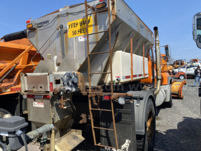 Truck-mounted snow plow in-yard showing dump hopper and rear mechanical components; exterior metal frame and hoses visible.