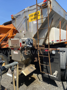 Truck-mounted plow truck displaying rear hopper with ladder and hydraulic components; visible label on yellow sign reading YEEP BACK 50 FEET; Mack branding on mudflap; metal framework and piping around hitch.