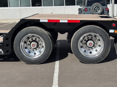 Trailer wheel assembly showing dual rear axles with polished rims and black frame, wooden deck above, parking lot background.