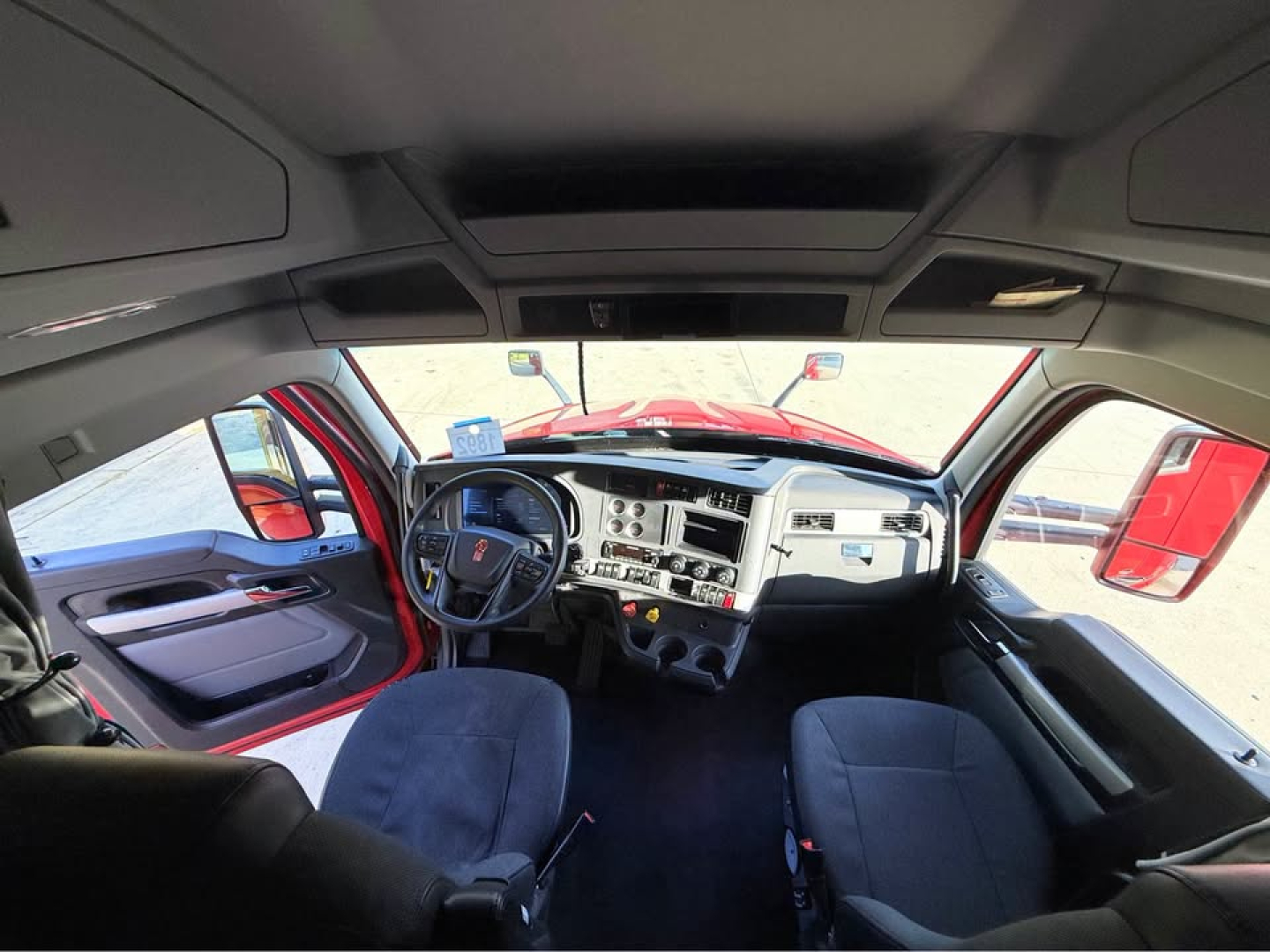 Cab interior of a Kenworth truck — steering wheel, dashboard with numerous gauges and controls, two front seats, overhead console. Text visible on dashboard includes standard truck controls but no legible model text.