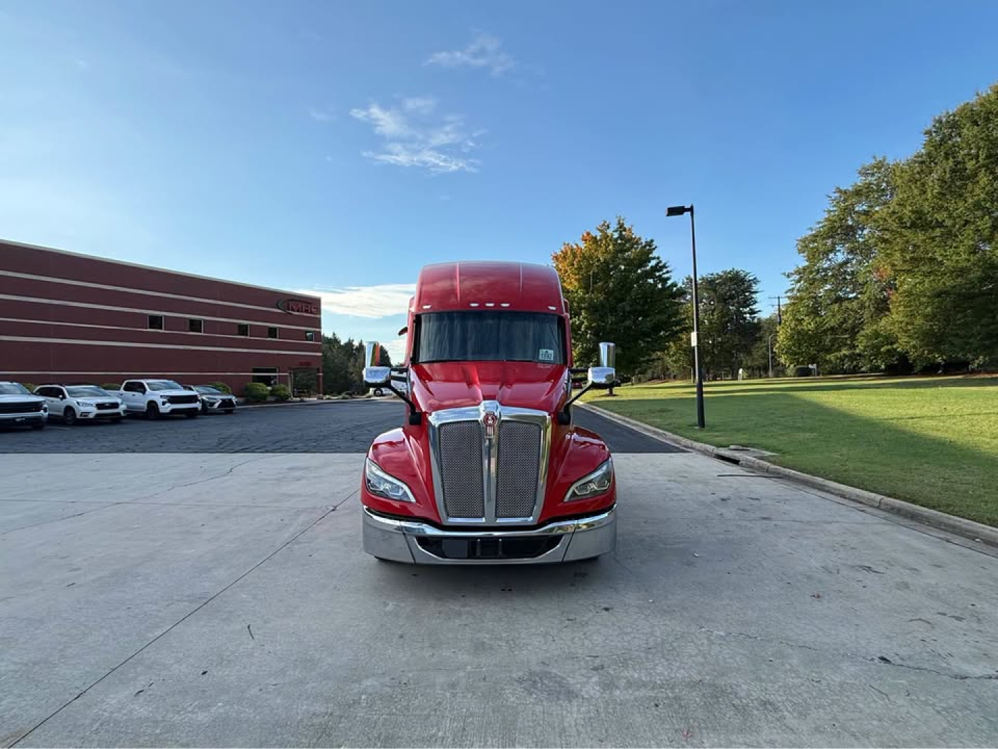 Kenworth T680 tractor front view in red with chrome grille and bumper, on a concrete lot with trees in the background.