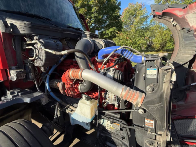 Kenworth T680 engine bay view showing red engine with blue silicone turbo hose, gray intake tube, black radiator shroud, and various hoses and reservoirs.