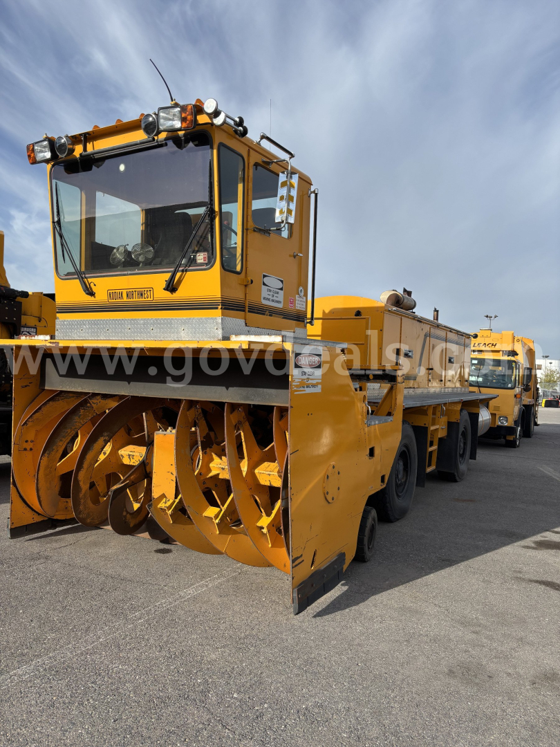 Snow plow truck with snow blower attachment on front, yellow cab and equipment body, Kodiak logo on cab window; multiple safety mirrors and lights; field with other yellow trucks in background.