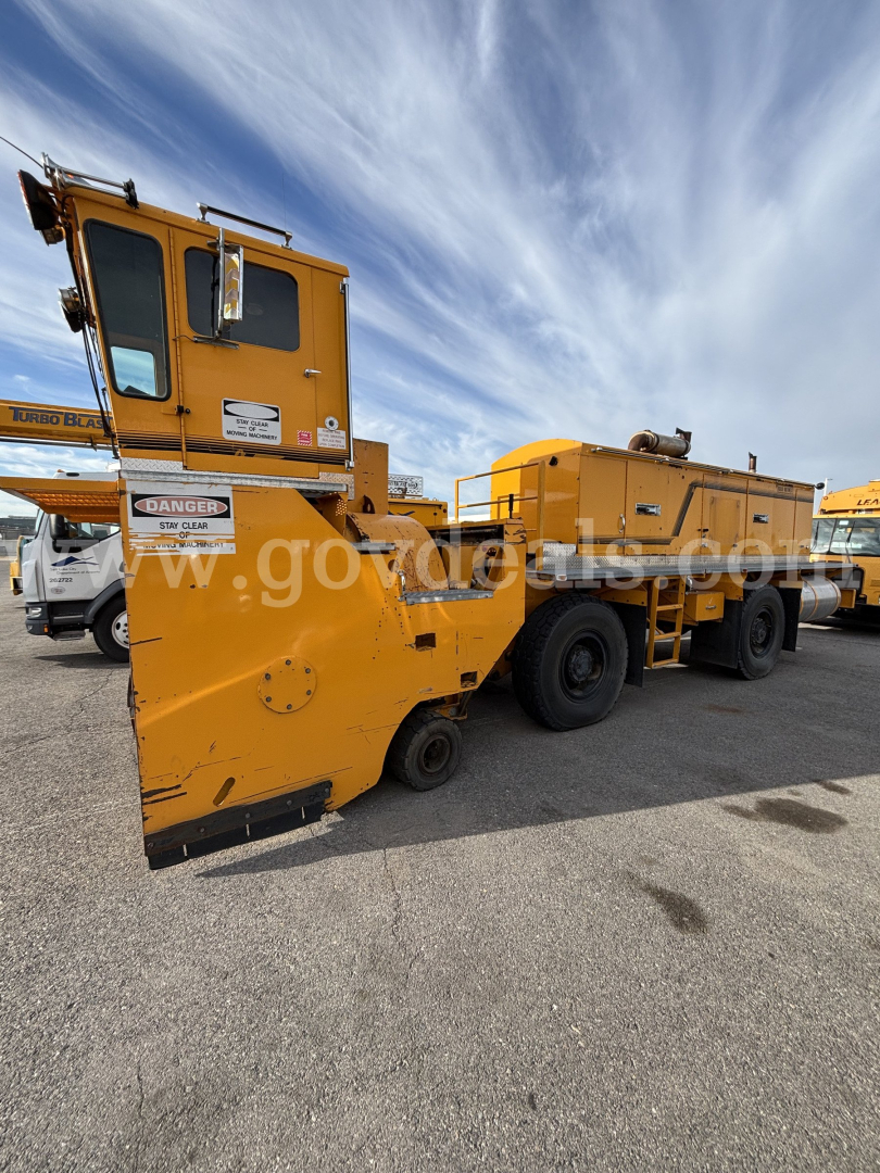 Yellow Kodiak CD6072-119 maintenance vehicle with large front plow and flatbed trailer in a parking lot.