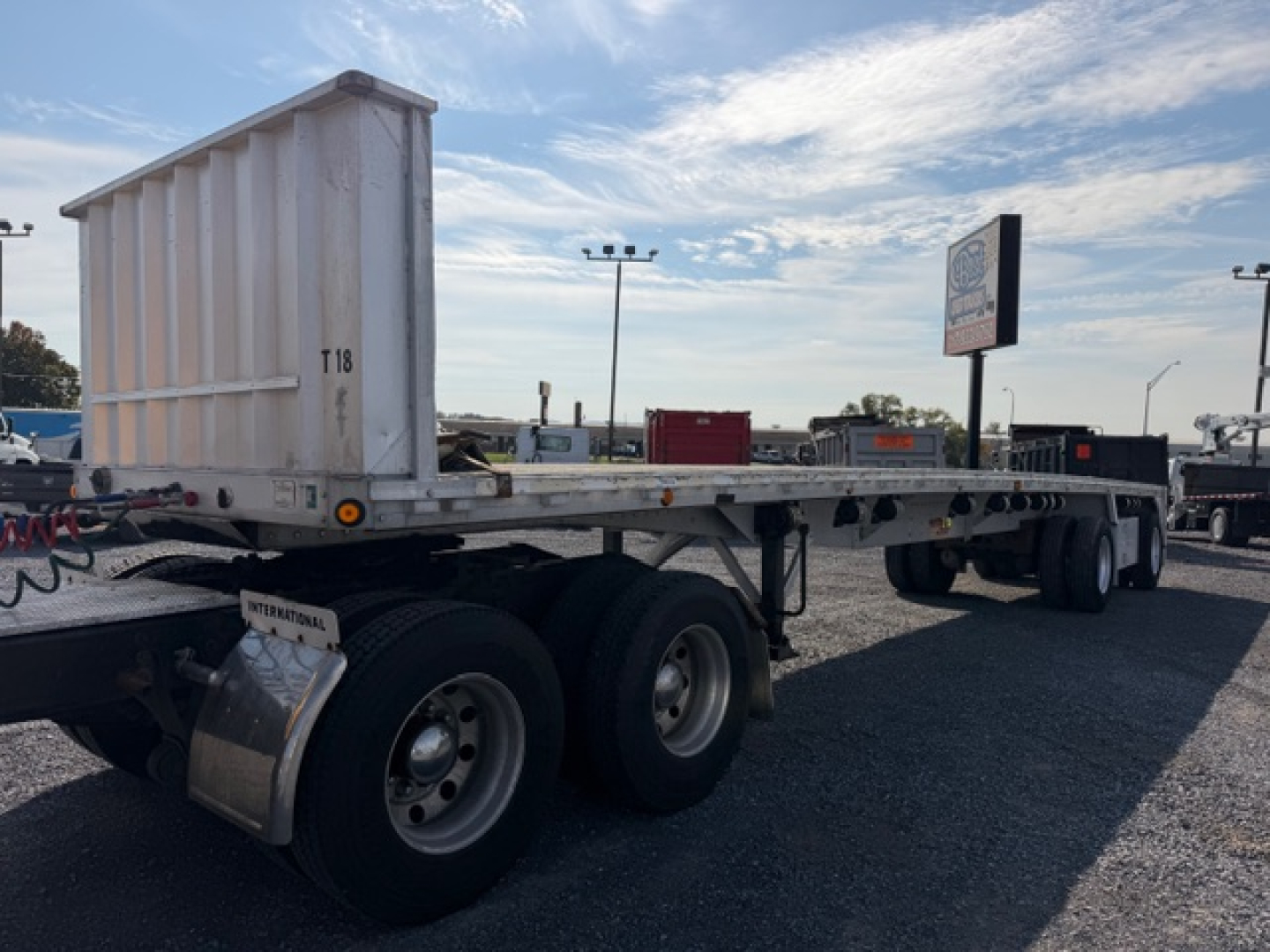Flatbed trailer showing dual rear axles and a white behind cab storage box with RAM-style bed. Visible text on sticker near wheel and in background.