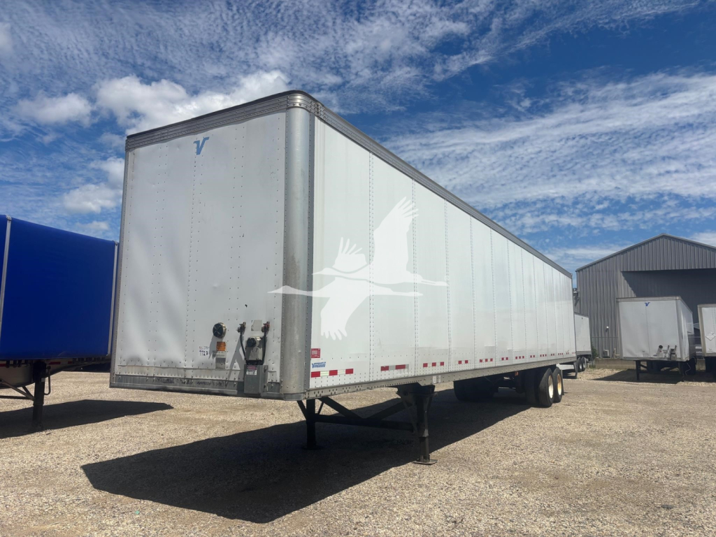 Trailer side view showing Vanguard logo on the front and a white trailer with a bird-like logo; twin rear wheels; gooseneck hitch area visible; blue sky with clouds.