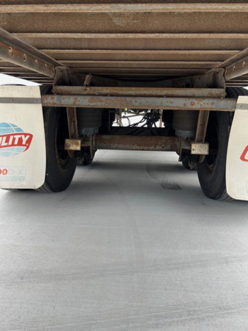 Rear view undercarriage of a utility trailer with wheels and axle, showing rusted steel frame, suspension components, and white mudflaps with red logoPartial text visible: 'UTILITY' on mudflap.