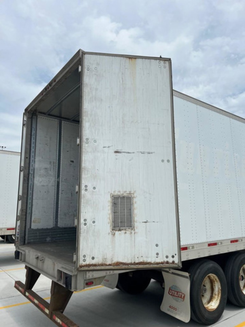 Utility van trailer showing rear lift gate area and open cargo door with exposed interior walls and wheel well. Visible cement ground and other trailer in background.