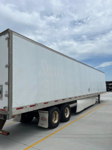 Trailer side view showing a long white box trailer with triple axle and wheel wells, no visible branding