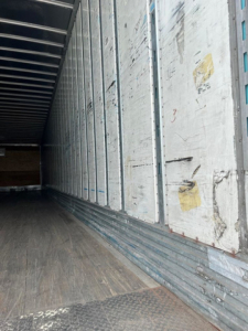 Trailer interior showing metal side walls with vertical seams and a textured wooden floor; long, empty cargo area.