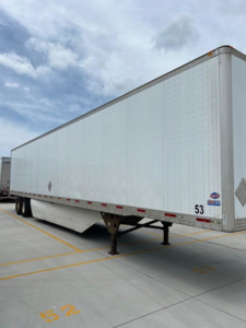Utility van trailer showing a long white rectangular cargo box on a raised landing gear with a black undercarriage, blue sky with clouds.