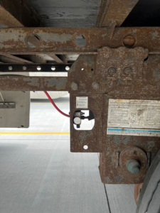 Trailer hitch and rusty undercarriage with identification sticker and bolt holes; side of a utility van trailer showing rusted metal frame and a small white label.