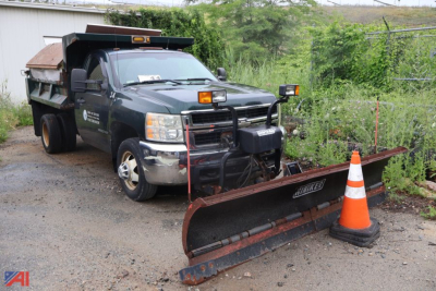 Chevrolet Silverado 3500HD dump truck with plow and salt/sander setup, front grille guard, orange cone, metal snow plow blade attached, dark green body, gravel lot.