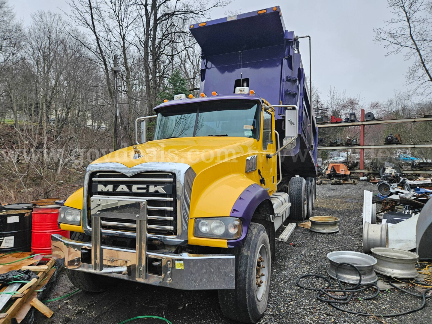 2014 Mack GU713 dump truck with yellow cab and purple dump bed, front grille with MACK emblem, exposed front bumper guard, in a salvage lot with spare parts and trees in background.