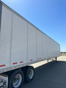 Trailer side view showing a white Wabash van trailer with tandem axles and red reflective tape along the lower edge.