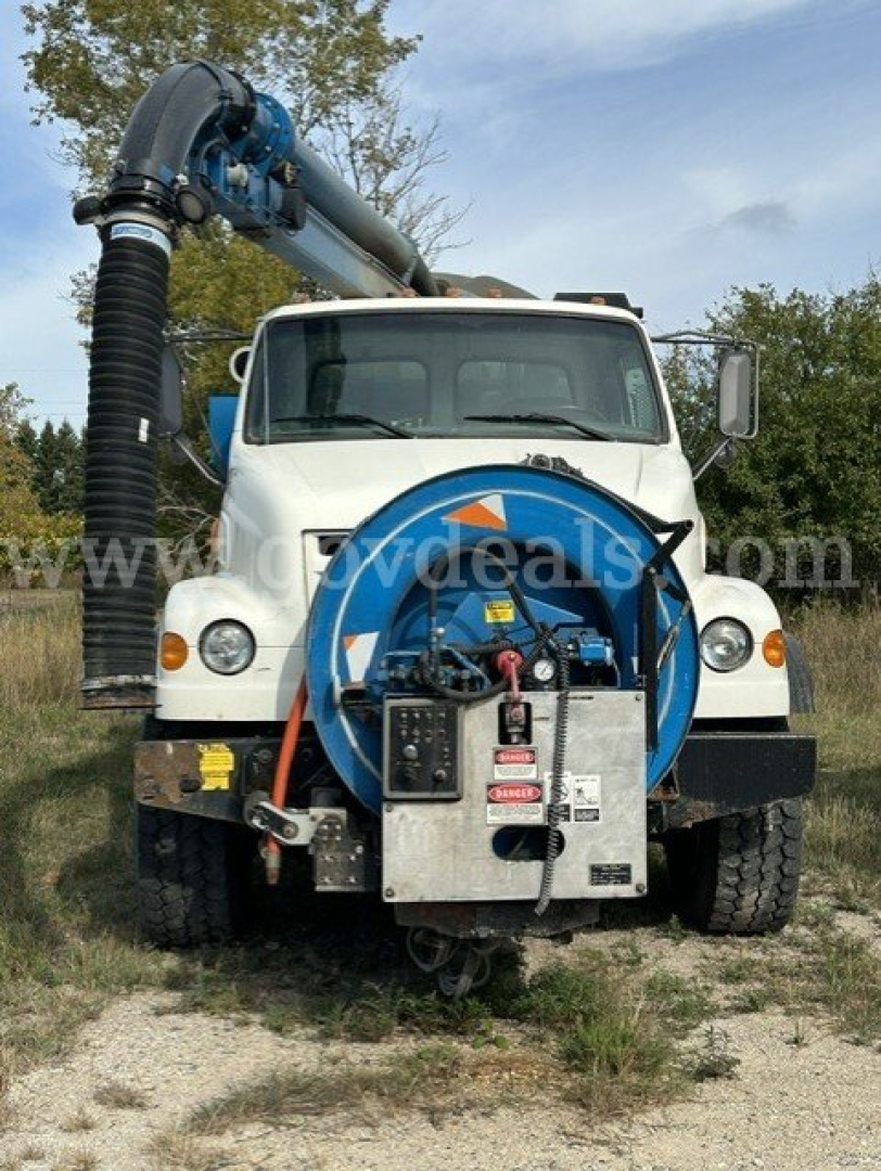 Front view of a white Sterling L7501 vacuum truck with a blue hose reel and black exhaust/vacuum arm mounted on the back, visible on a grassy field.