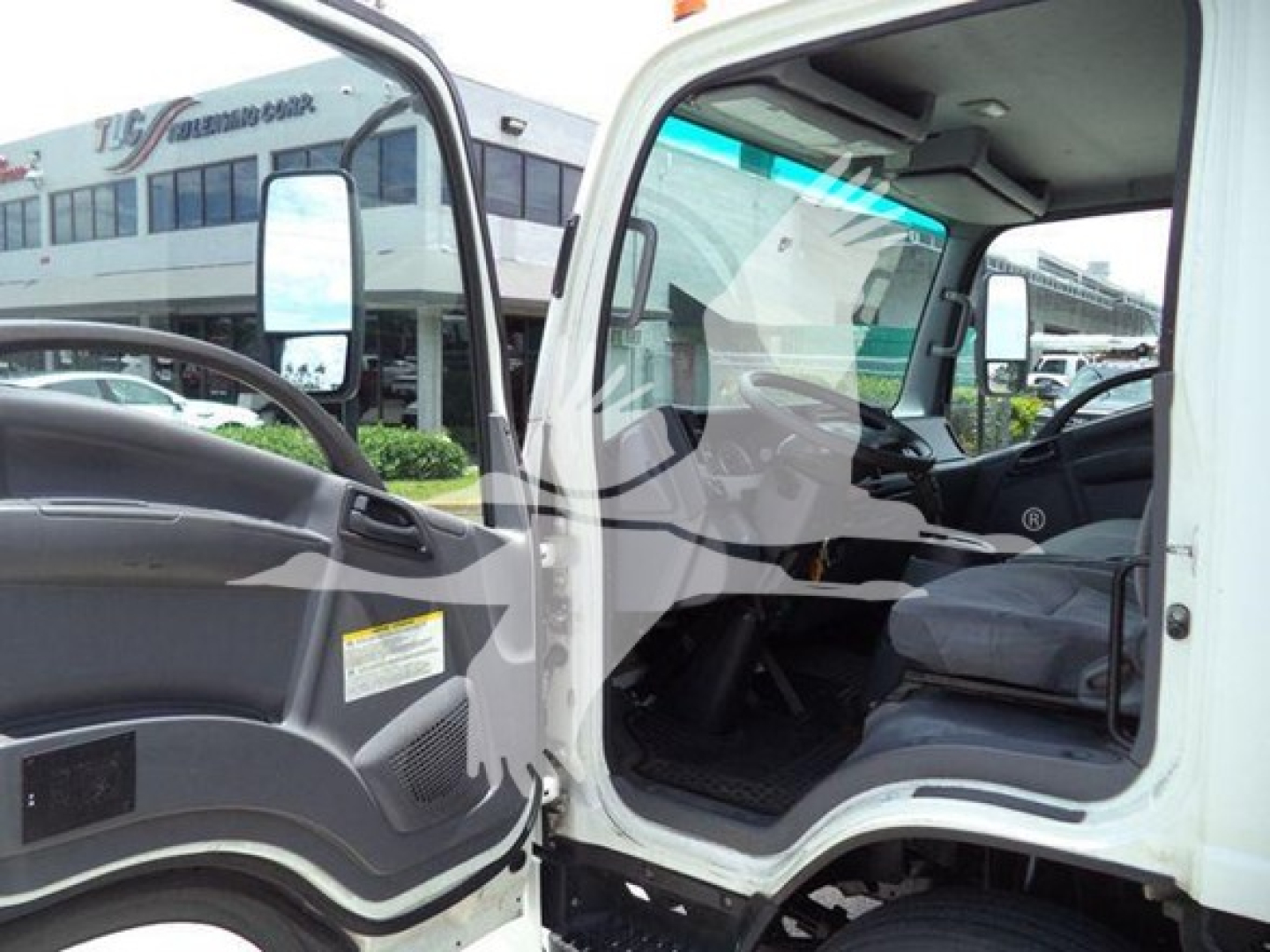Isuzu NRR truck cab interior and open door showing steering wheel and driver seat with dashboard, exterior side mirror, and view of box truck in background.