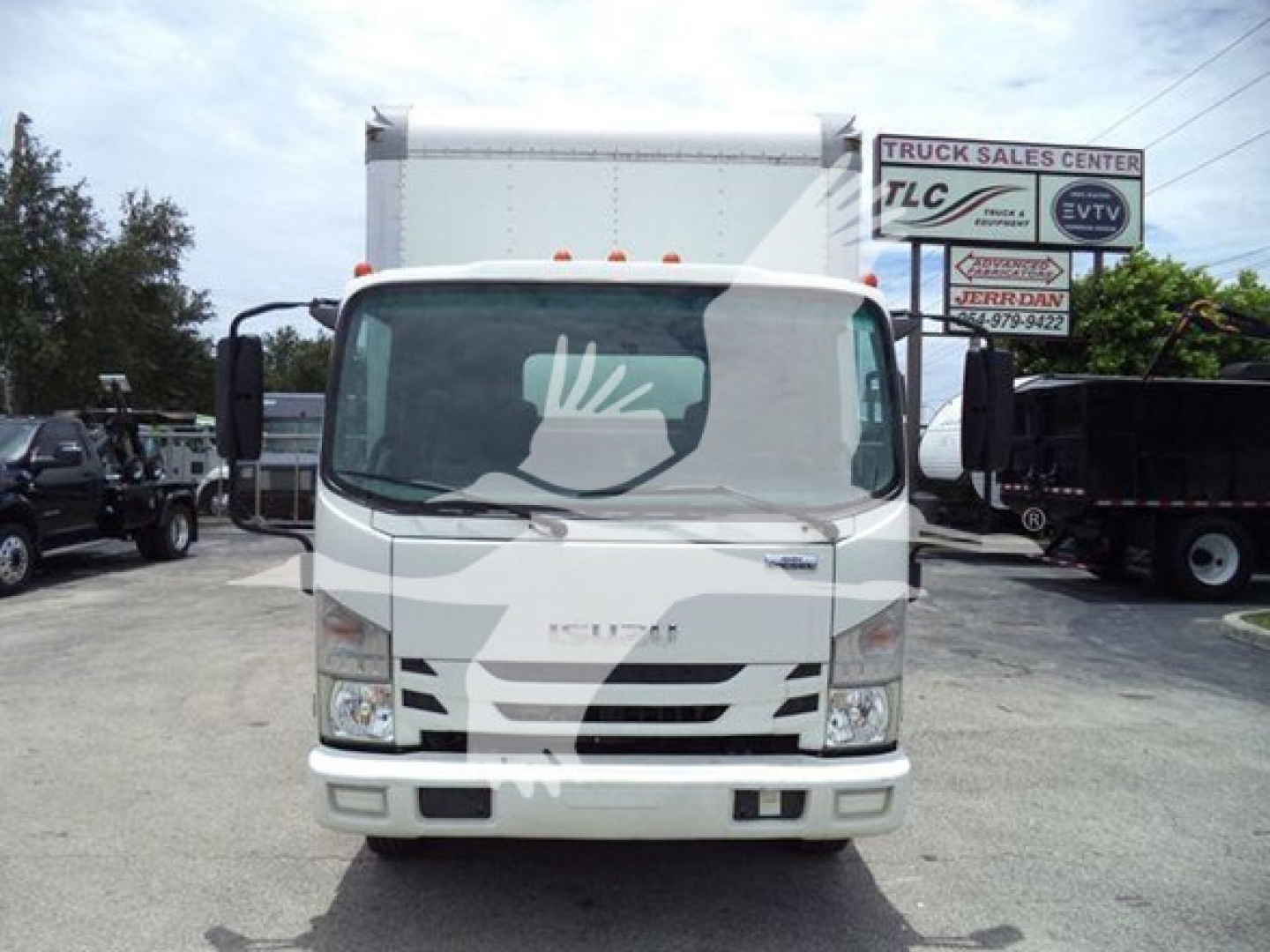 Isuzu NRR box truck front view with stock signs in background. Visible text includes 'ISUZU' on grille and some signs on right side of lot. No other legible model or specification text clearly readable in the image.