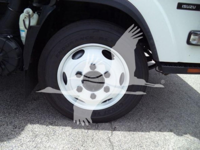 Isuzu NRR front wheel and fender showing wheel with white hubcap and tire, with partial Isuzu badge on the body and a coolant reservoir visible in engine compartment.