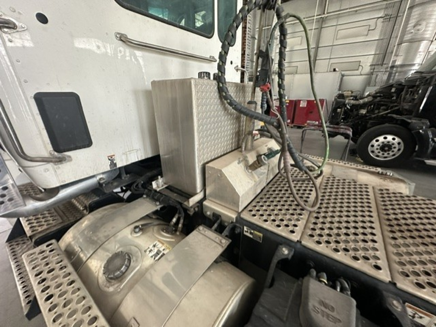 Kenworth T880 truck cab area showing aluminum fuel tank and platform with diamond plate, hoses, and metal steps; partial engine bay and rear cab corner visible.