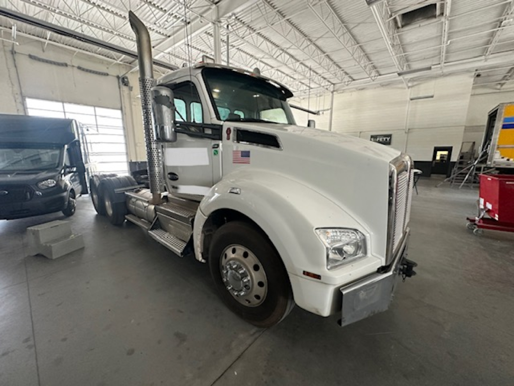 Kenworth T880 conventional daycab truck in showroom, front-left view, white cab with large exhaust stacks, chrome mirrors, and large bumper.