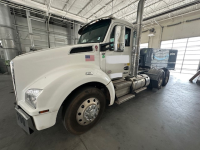 Kenworth T880 conventional daycab truck in a showroom with white cab and chrome exhaust stack, side view showing US flag decal and chrome steps.
