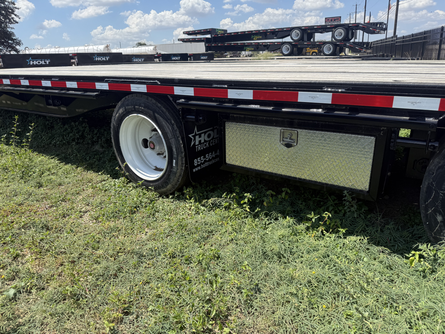 Flatbed trailer with black deck and white wheel, undercarriage toolbox and reflective red-white side rails visible.