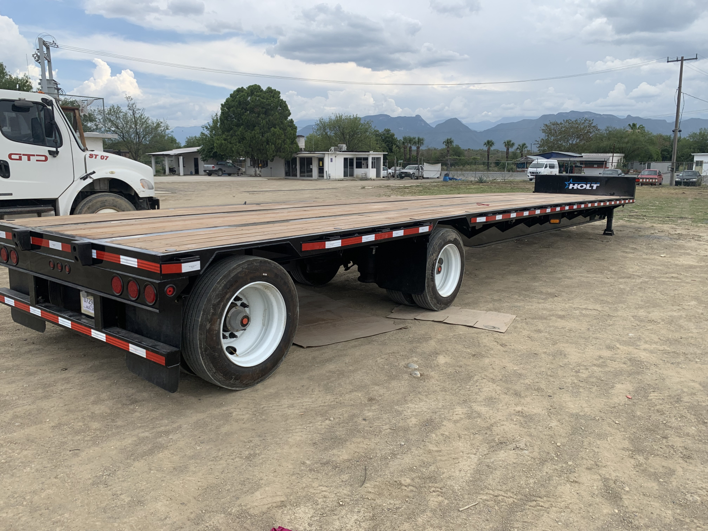 Flatbed trailer with wooden deck and dual rear axles, black frame, white rims, and red/white reflective tape. Visible text includes 'HOLT' on the front fender and a white truck in background.