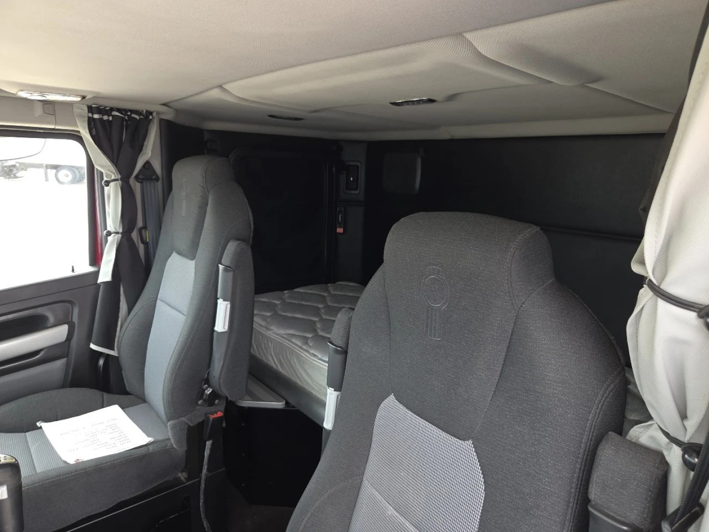Kenworth W990 sleeper cab interior showing driver seat and bunk area with gray fabric seats and overhead cab, dashboard not visible.