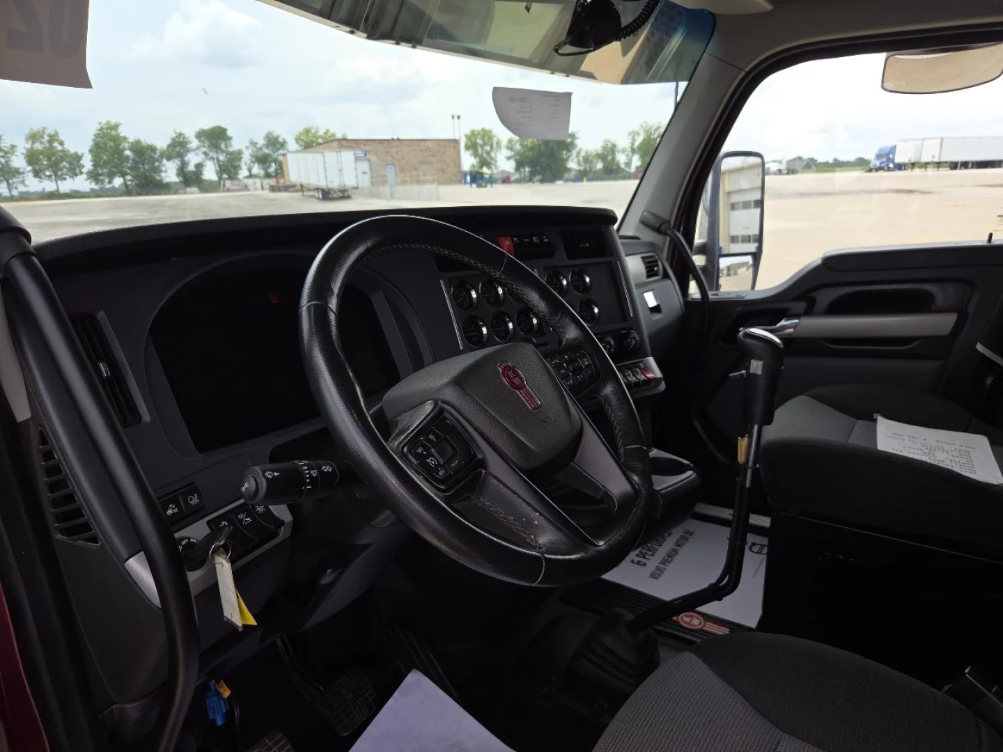 Kenworth W990 interior view showing steering wheel with red Kenworth emblem and dashboard gauges; gear shifter in center, driver seat, window, and outside yard visible.