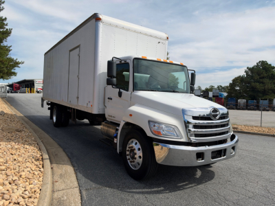 2019 Hino 268A box truck in a dealership lot, white cab with large box body, front grille and headlights visible, VIN tag partially legible.