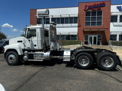 Truck side view of a 2022 Mack PI64T conventional daycab in front of a dealership building, showing a long flatbed with multiple axles and chrome fuel tanks.