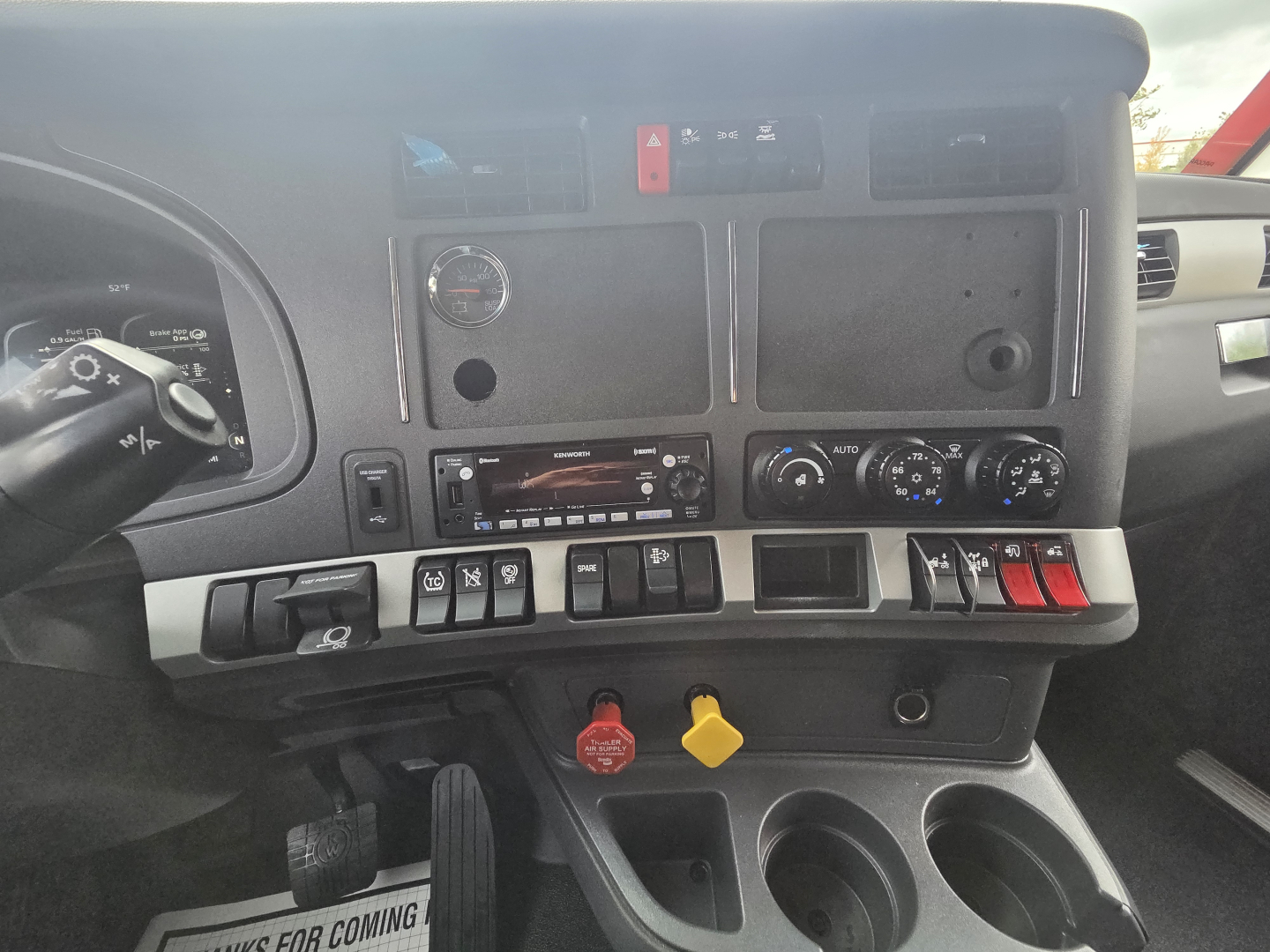 Dashboard area of a Kenworth truck showing various controls, switches, gauges, two cup holders, ignition area, and yellow and red latches. Visible text includes vehicle controls and blank panels.