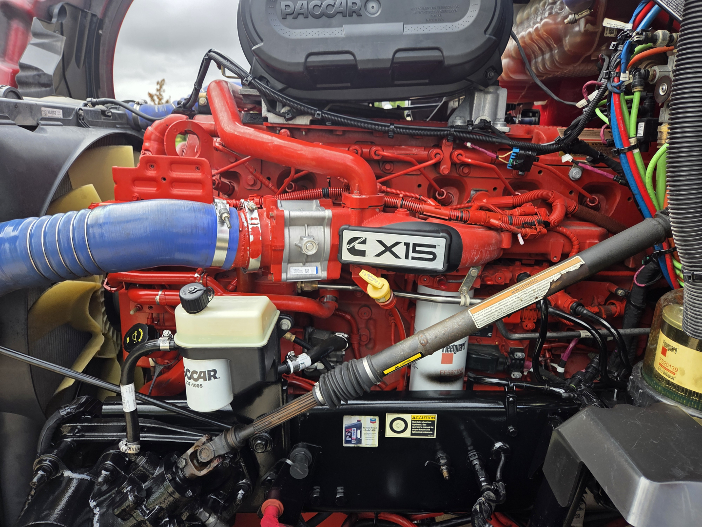 Kenworth T680 engine bay showing X15 engine block with red paint, blue hose, and various hoses and wiring.