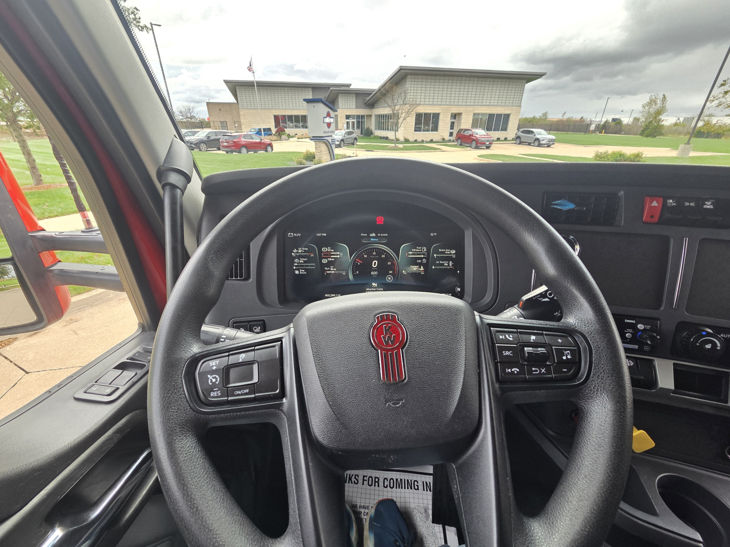 Kenworth truck interior steering wheel with dashboard gauges visible; various controls on steering wheel; center console; side window and outside view of dealership.
