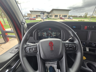Kenworth truck interior steering wheel with dashboard gauges visible; various controls on steering wheel; center console; side window and outside view of dealership.