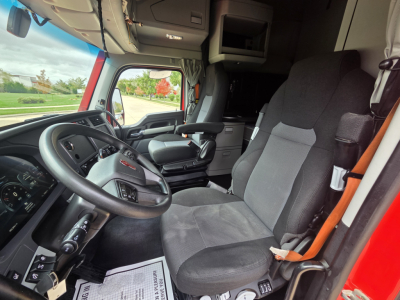 Interior of a Kenworth truck cabin showing steering wheel, driver seat, and dashboard with visible controls; gray fabric seats; open door to outside with red exterior.