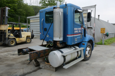 Truck cab view showing blue Freightliner CL112 daycab with cylindrical sleeper fuel tanks and vertical exhaust, cargo yard background.