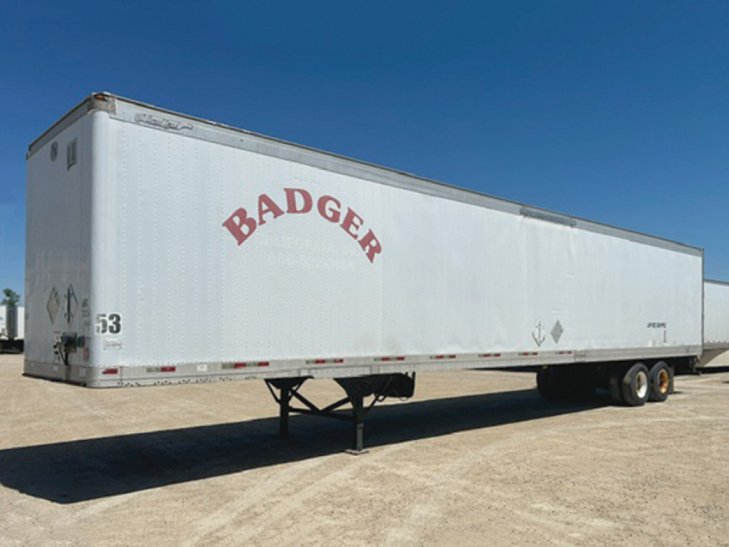 Trailer with 'BADGER' lettering on side, long white cargo trailer in a lot, dual rear wheels, front support jack under the hitch.
