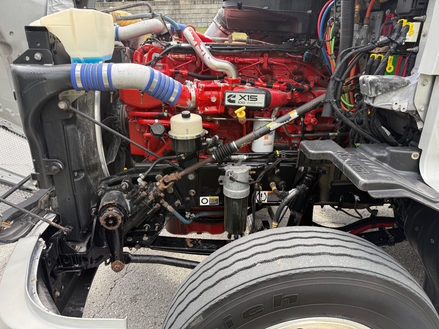 Kenworth T680 engine bay showing 15-liter inline-6 diesel engine with red paint, blue silicone hose, coolant reservoir, and various hoses and electrical wires.