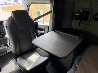 Kenworth T680 cab interior showing driver's seat, swivel table, and sleeper area with mounting bracket. Dashboard and windshield visible in background.