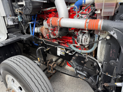 Kenworth T680 engine bay with bright red engine block, silver exhaust pipes, and various hoses; front left wheel and suspension visible.