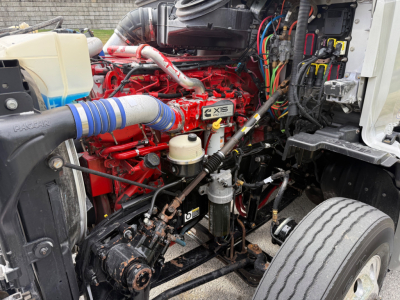 Kenworth T680 engine bay with red engine, blue intercooler hose, various hoses and wiring, fuel filters, and metal chassis visible.