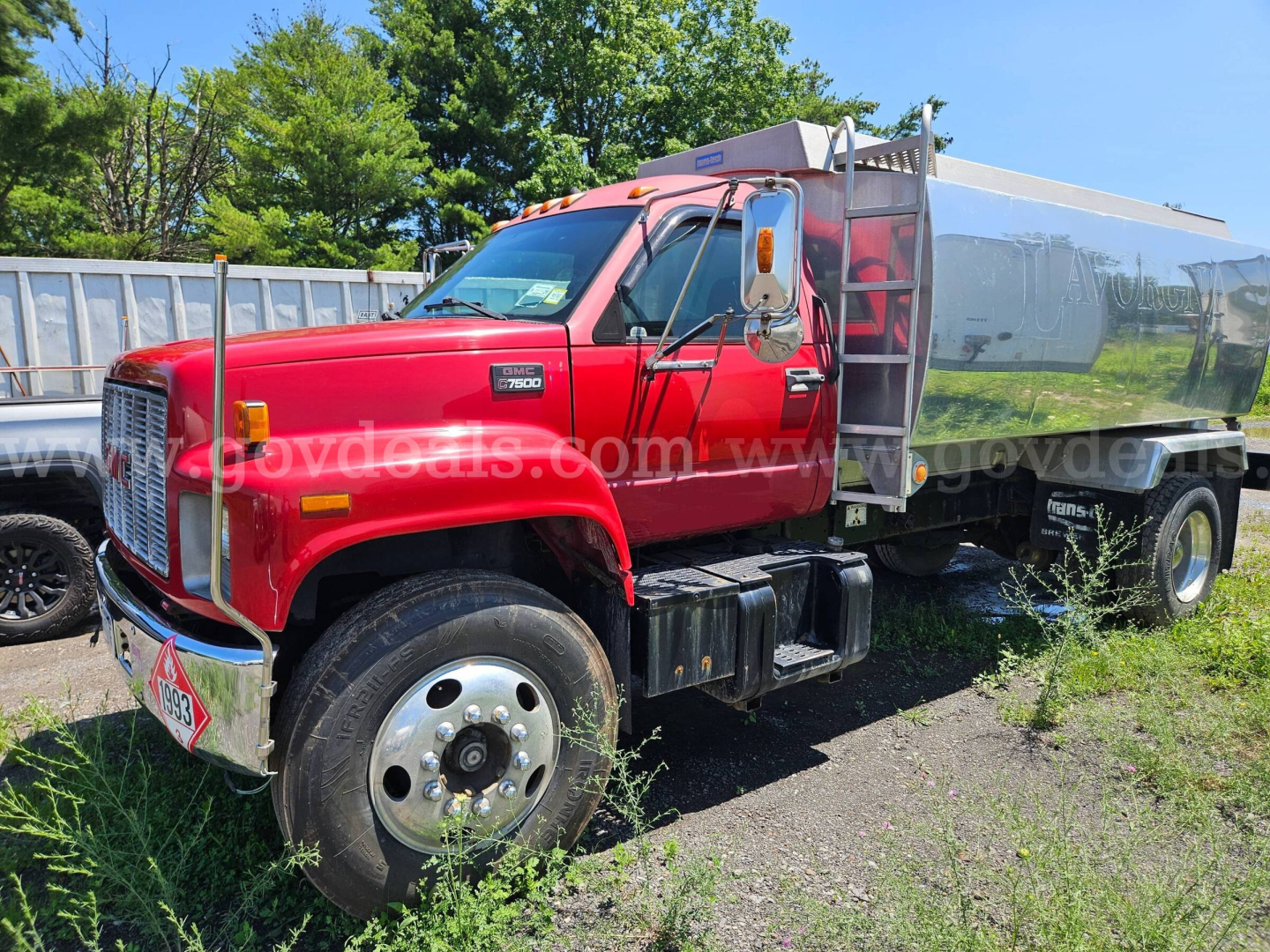 GMC C7500 dump truck in red with fuel/tank body, front side view showing large chrome grille, single rear axle, and polished steel wheels.
